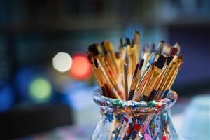 Clear jar covered with paint splatters, holding a variety of small paint brushes.